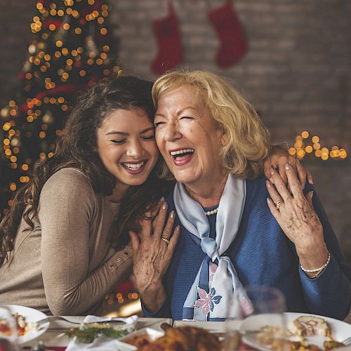Two women share a joyful moment at a festive dinner table, with a decorated Christmas tree and stockings in the background.