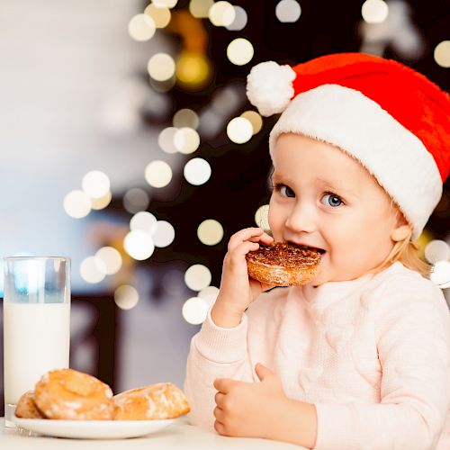 A child in a Santa hat enjoys a donut with milk beside a Christmas tree, glowing with lights in the background.