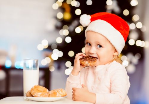 A child in a Santa hat is eating a donut with a plate of donuts and a glass of milk in front of a Christmas tree ending the sentence.
