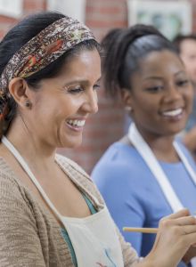 Two women are smiling and painting while wearing aprons in a creative studio setting.