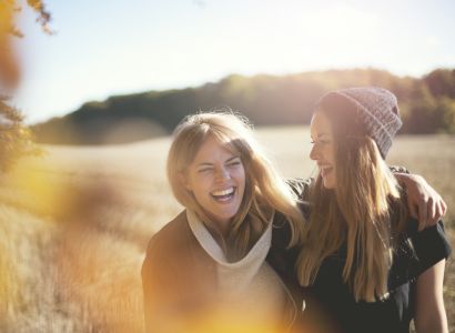 Two people are laughing and walking together in a sunny outdoor setting, with trees and open fields in the background.