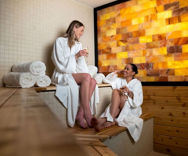 Two people in white robes enjoy a relaxing moment in a sauna, sitting on wooden benches with drinks, against a glowing amber wall.
