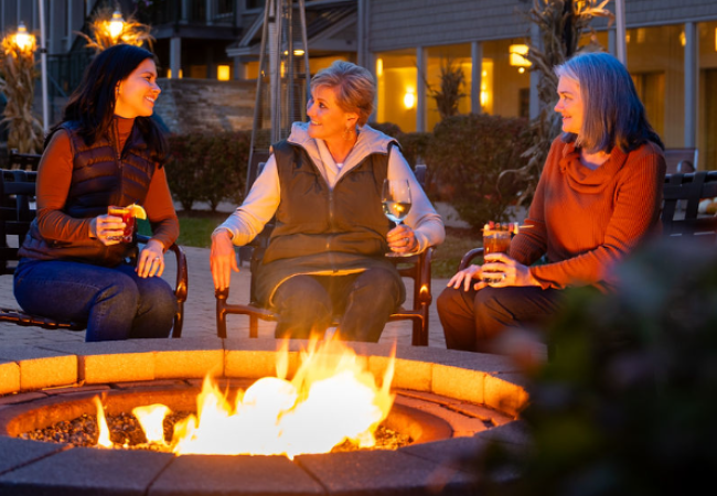 Three people sit around a fire pit outside a building, with string lights above them, enjoying drinks.