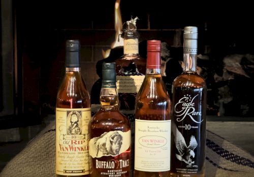 Five bottles of bourbon, including brands like Buffalo Trace and Pappy Van Winkle, are displayed in front of a fire.