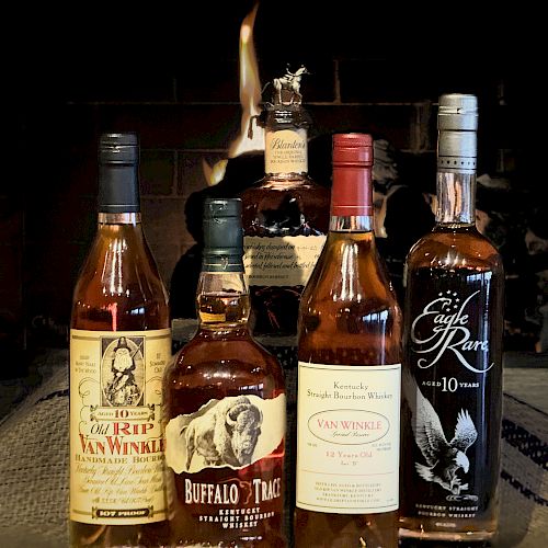 Five bottles of bourbon, including brands like Buffalo Trace and Pappy Van Winkle, are displayed in front of a fire.