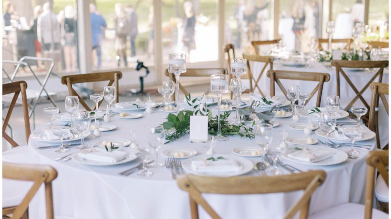 A round table set for a formal event with white tablecloth, greenery centerpiece, glassware, and wooden chairs.