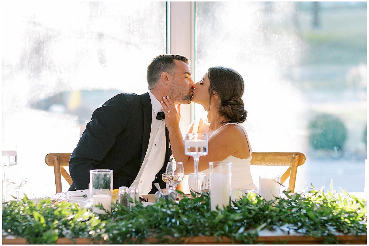 A couple at a wedding reception shares a kiss at a decorated table with candles and greenery in front of a bright window.