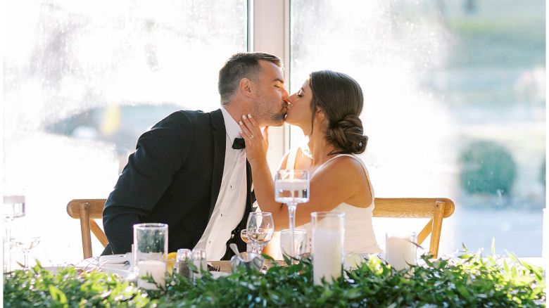 A couple at a wedding reception shares a kiss at a decorated table with candles and greenery in front of a bright window.