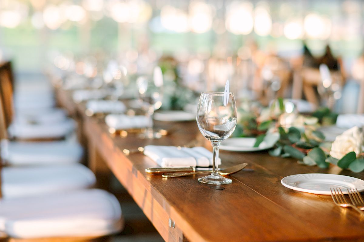 A long wooden table is set for a formal event, with white plates, silverware, wine glasses, and floral decorations.