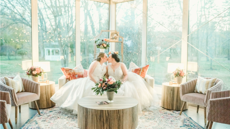 Two brides in white dresses share a kiss in a beautifully decorated room with large windows, cozy seating, and floral arrangements.