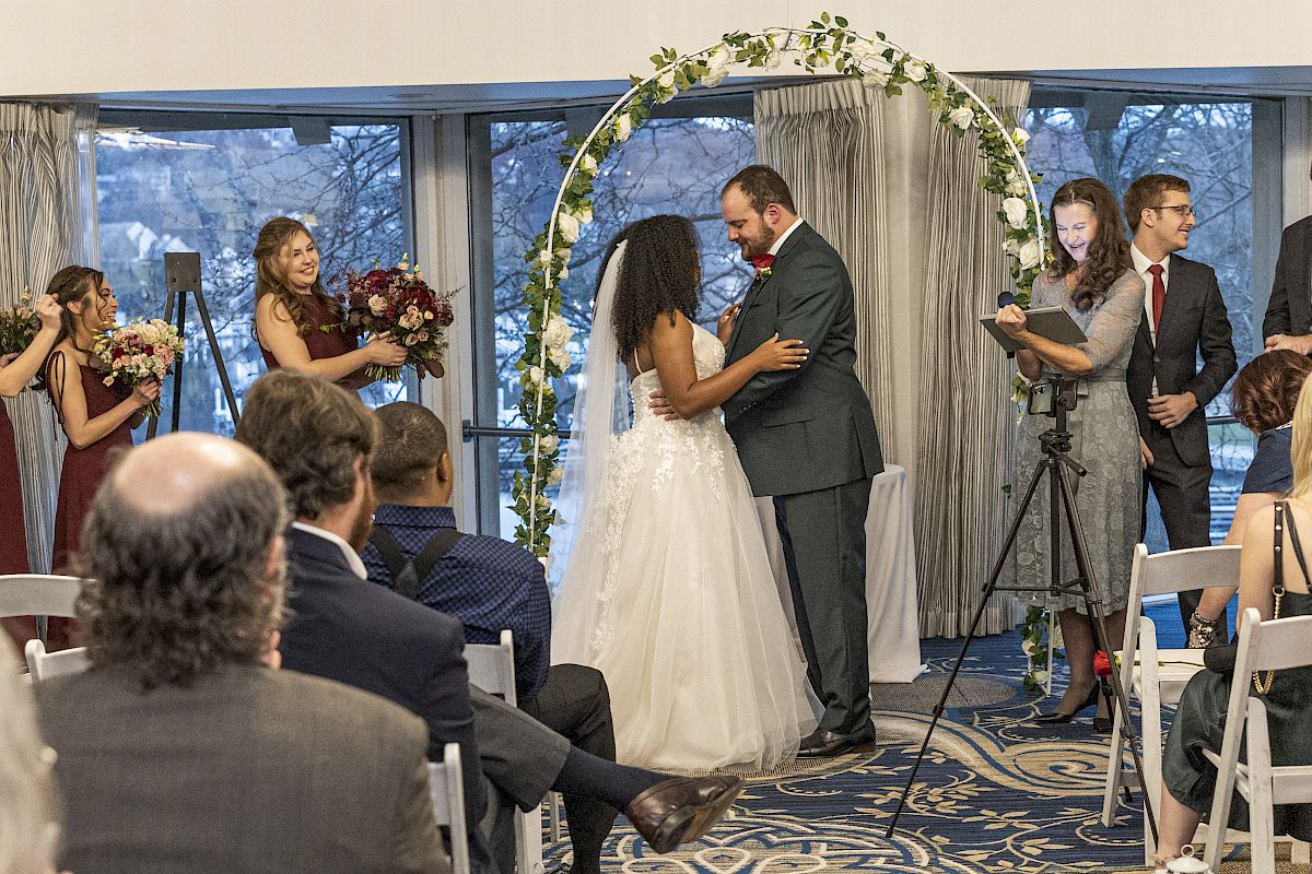 A couple is at the altar in a wedding ceremony, surrounded by their wedding party and guests, under a decorated arch while an officiant oversees.
