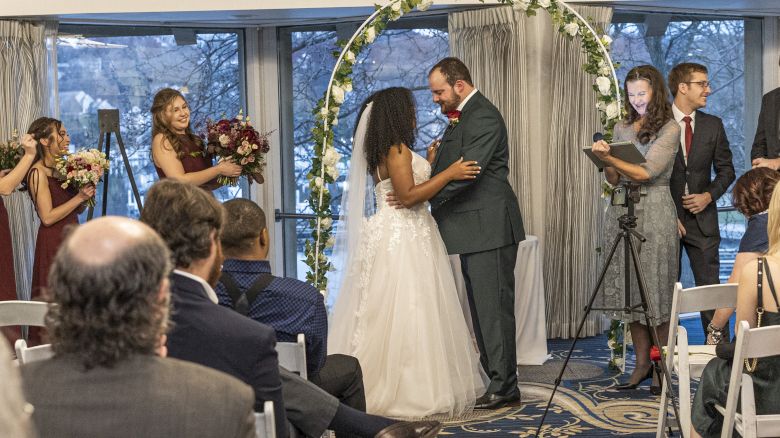 A couple is at the altar in a wedding ceremony, surrounded by their wedding party and guests, under a decorated arch while an officiant oversees.