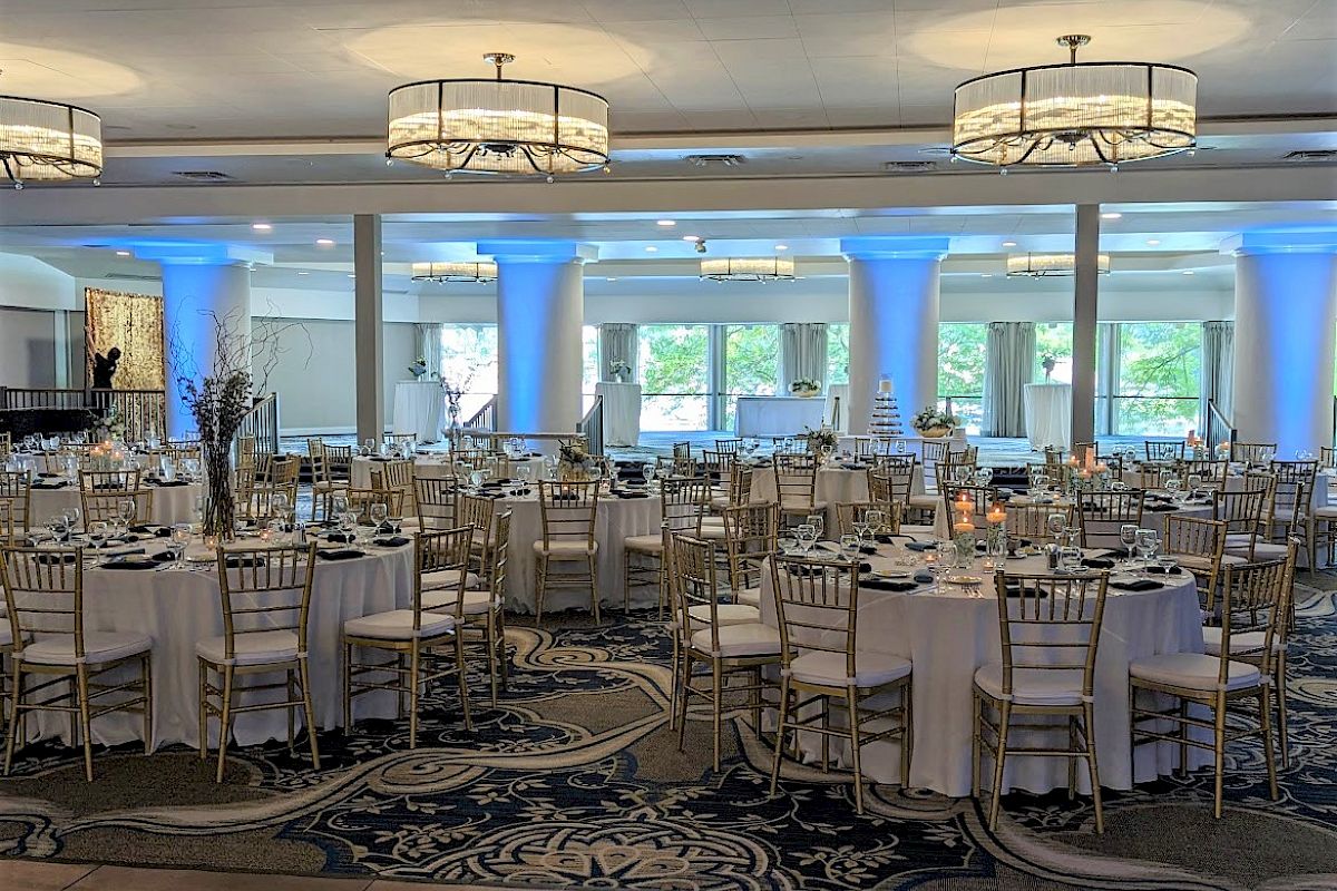 A large, elegantly decorated banquet hall with round tables, white tablecloths, and gold chairs prepared for an event with blue uplighting.