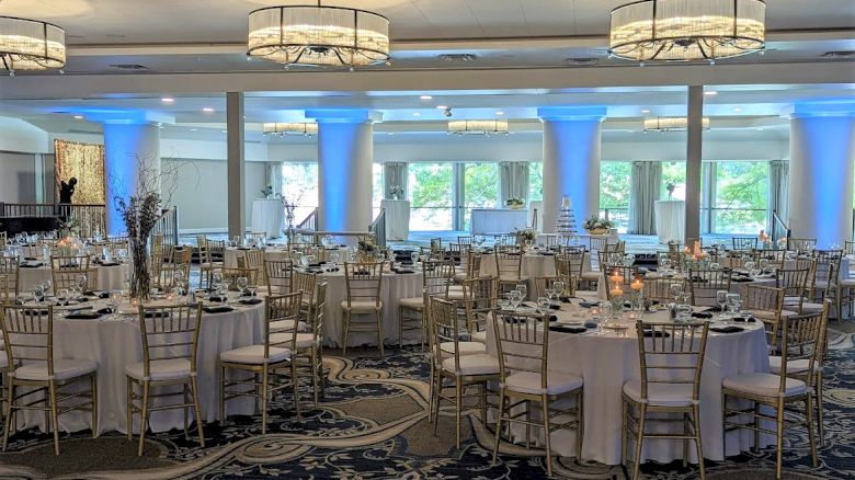 A large, elegantly decorated banquet hall with round tables, white tablecloths, and gold chairs prepared for an event with blue uplighting.