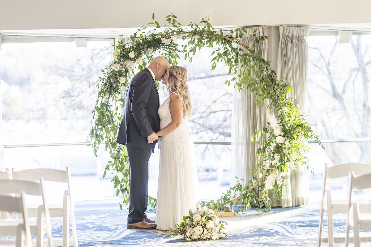A couple in formal attire is standing under a circular floral arch, holding hands and touching foreheads, in a bright, airy setting.