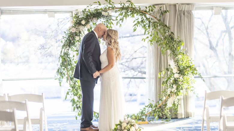 A couple in formal attire is standing under a circular floral arch, holding hands and touching foreheads, in a bright, airy setting.