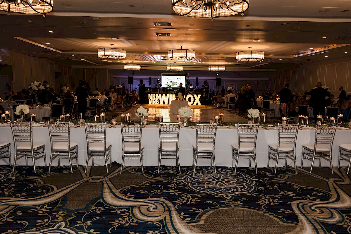 A decorated event hall with long tables, white chairs, chandeliers, and a lit 