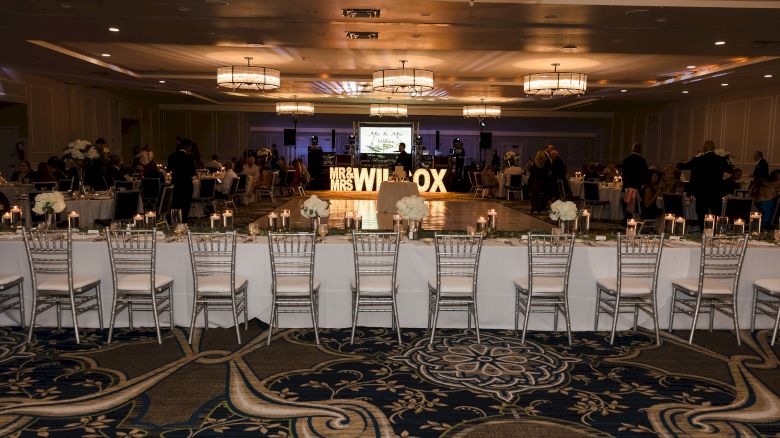 A decorated event hall with long tables, white chairs, chandeliers, and a lit 