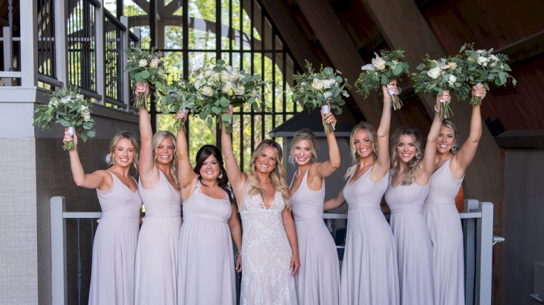 A bride stands center, surrounded by six bridesmaids in lavender dresses, all holding bouquets aloft, in an airy venue with natural light.