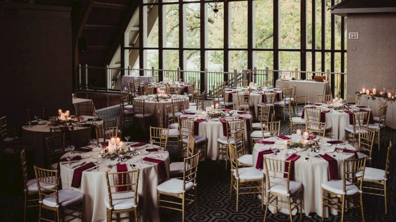 A decorated event space with round tables, white tablecloths, red napkins, and gold chairs, ready for a gathering or celebration, with candles on tables.