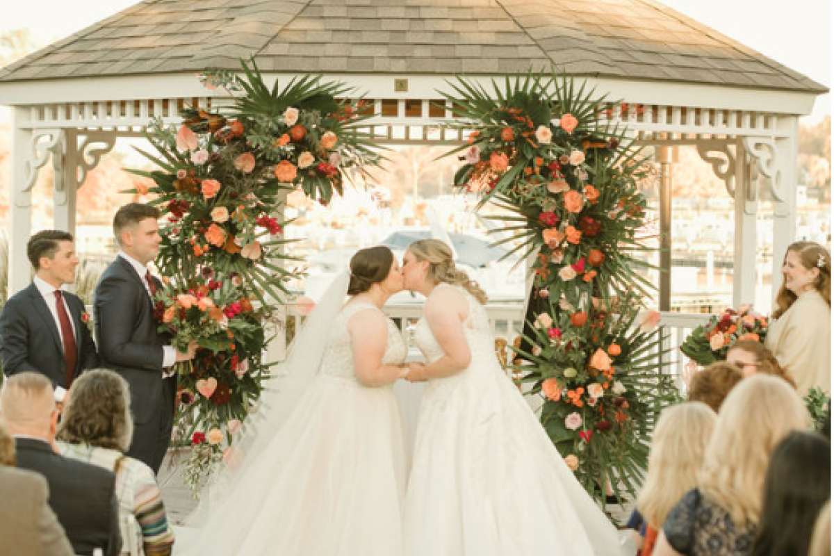 A wedding ceremony with two brides kissing under a gazebo adorned with flowers, attended by guests seated in white chairs.