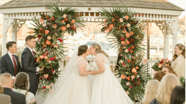 A wedding ceremony with two brides kissing under a gazebo adorned with flowers, attended by guests seated in white chairs.