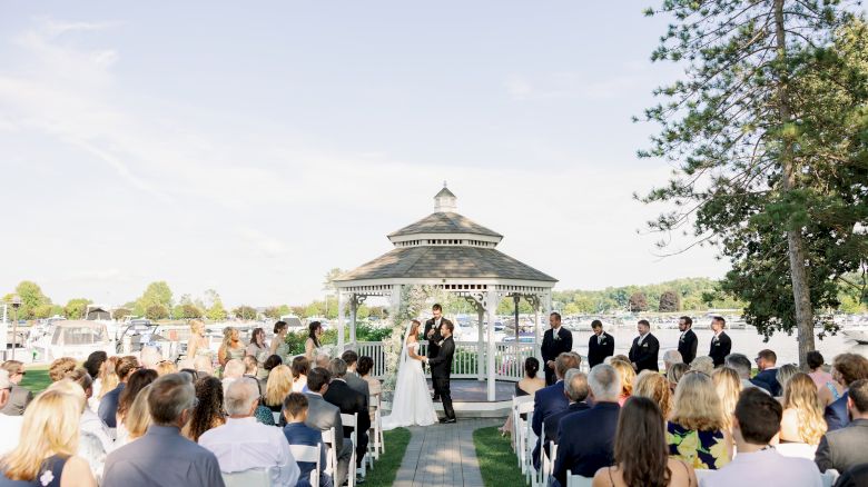 A wedding ceremony in progress outdoors by a gazebo with a crowd seated on both sides, the couple exchanging vows in the center.
