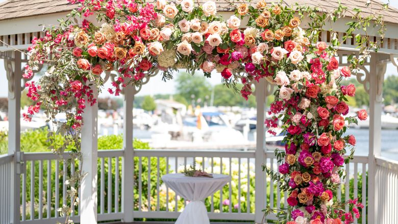 A decorated gazebo with a covering of colorful flowers, possibly set up for a wedding or an outdoor event, located near a body of water.
