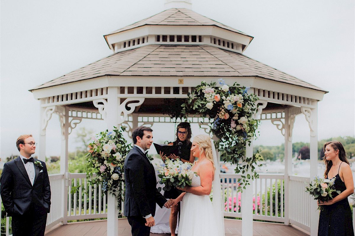 A couple is getting married in an outdoor gazebo, surrounded by greenery and flowers, with two attendants and an officiant present.