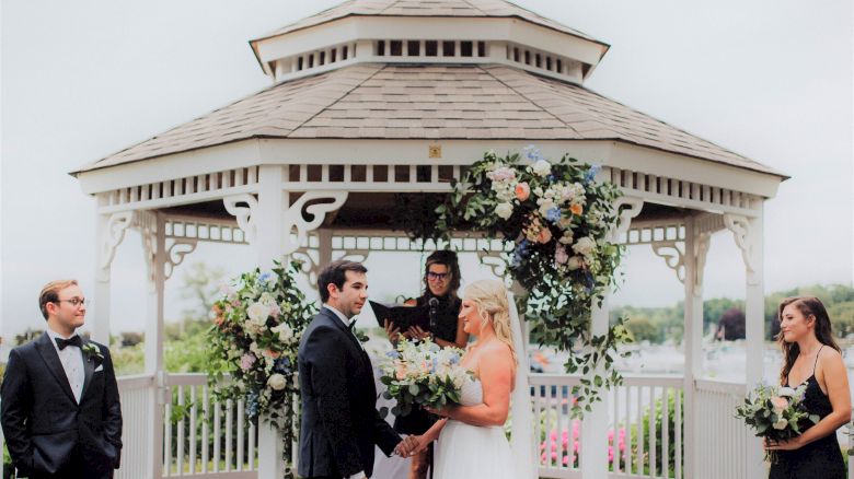 A couple is getting married in an outdoor gazebo, surrounded by greenery and flowers, with two attendants and an officiant present.