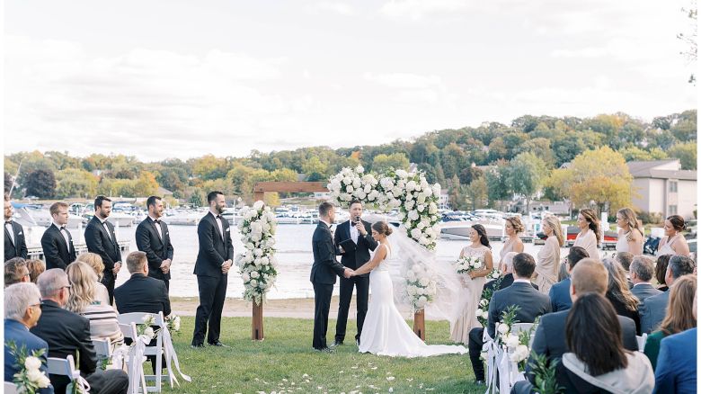 A wedding ceremony by the lake, with a couple under a floral arch, surrounded by guests and wedding party. Everyone is facing the couple.