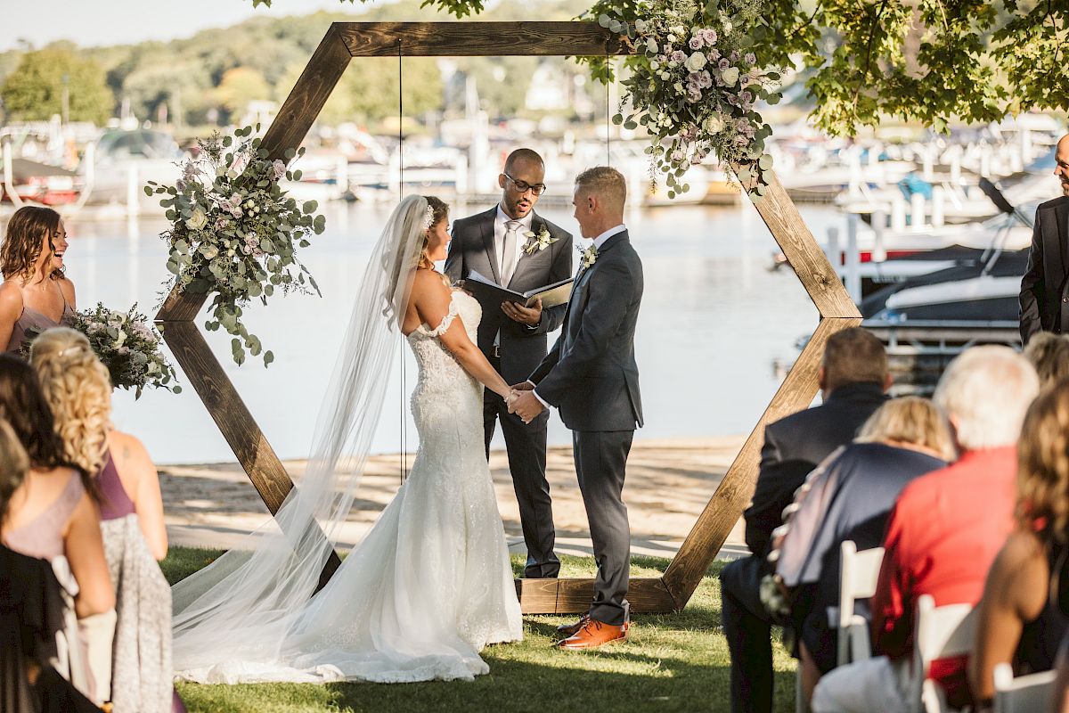 A couple is getting married outdoors by a lake, standing under a wooden hexagonal arch with flowers, as the officiant conducts the ceremony.