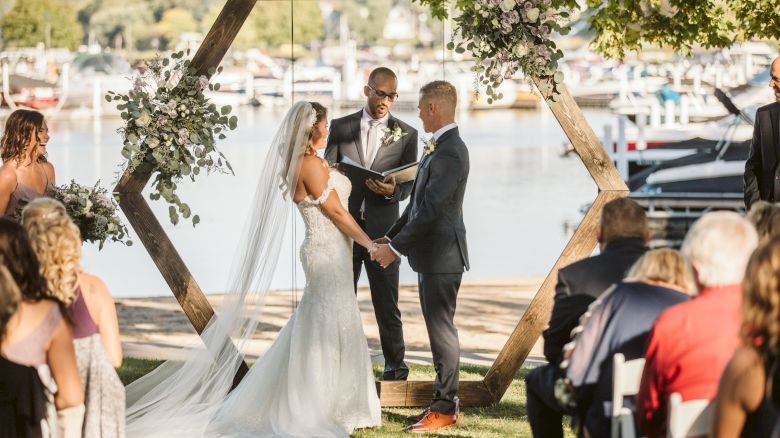 A couple is getting married outdoors by a lake, standing under a wooden hexagonal arch with flowers, as the officiant conducts the ceremony.