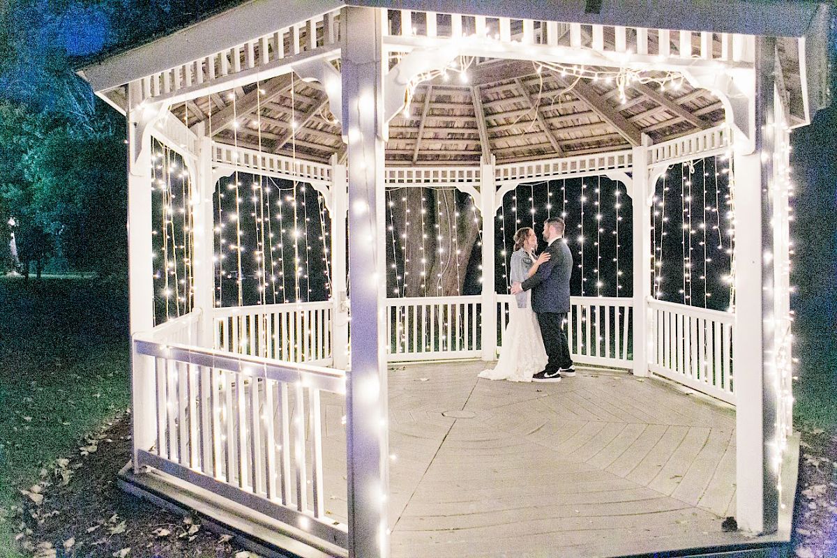 A couple is standing in a well-lit gazebo decorated with string lights, set against a dark outdoor background.