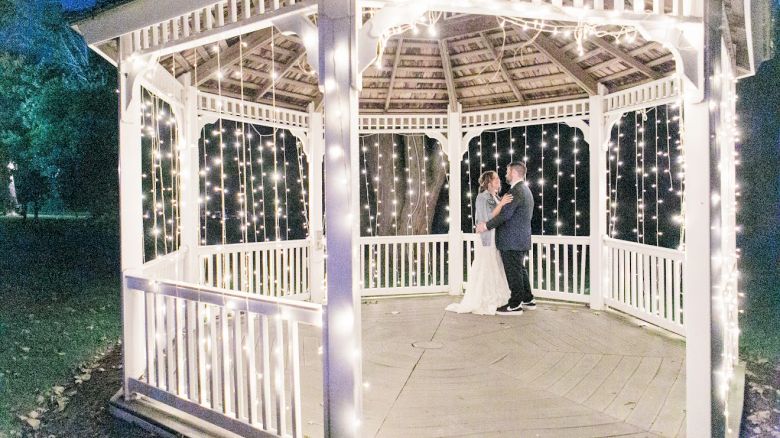 A couple is standing in a well-lit gazebo decorated with string lights, set against a dark outdoor background.