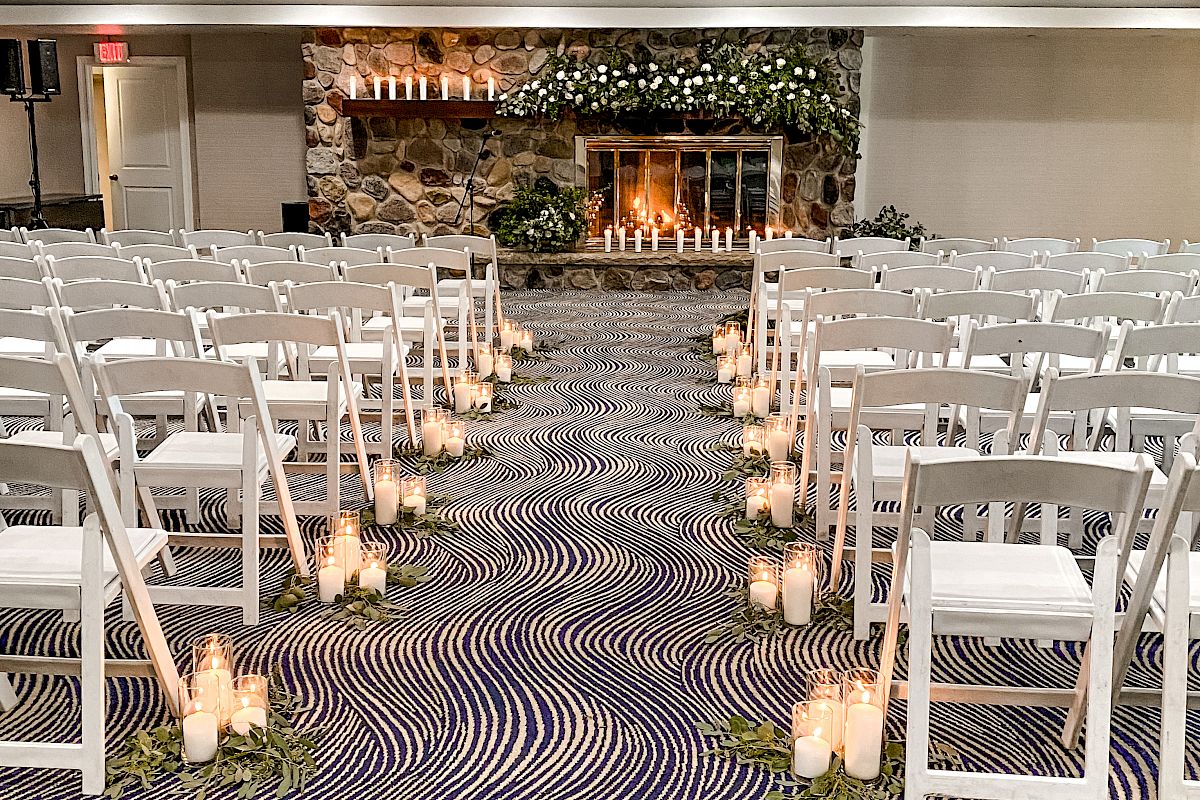 The image depicts a wedding ceremony setup with white folding chairs arranged in rows, candlelit aisle decor, and a stone fireplace adorned with greenery.