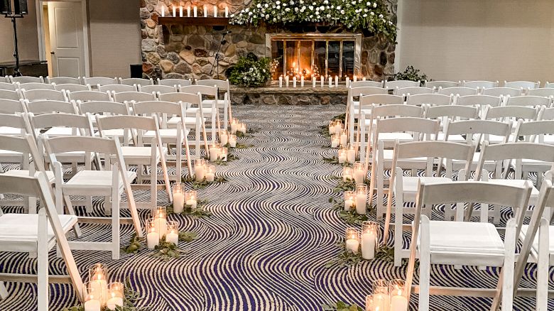 The image depicts a wedding ceremony setup with white folding chairs arranged in rows, candlelit aisle decor, and a stone fireplace adorned with greenery.