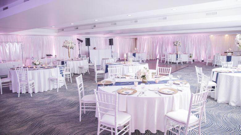 An elegantly set up event space with round tables, white chairs, centerpieces, and ambient lighting. The room is prepared for a wedding or banquet.