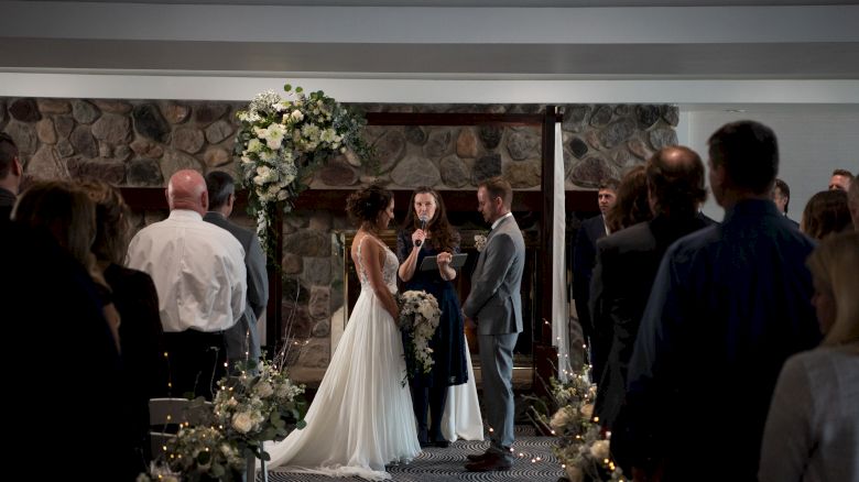 A wedding ceremony with a bride and groom facing each other, officiant in the middle, and guests seated on both sides in a decorated indoor venue.