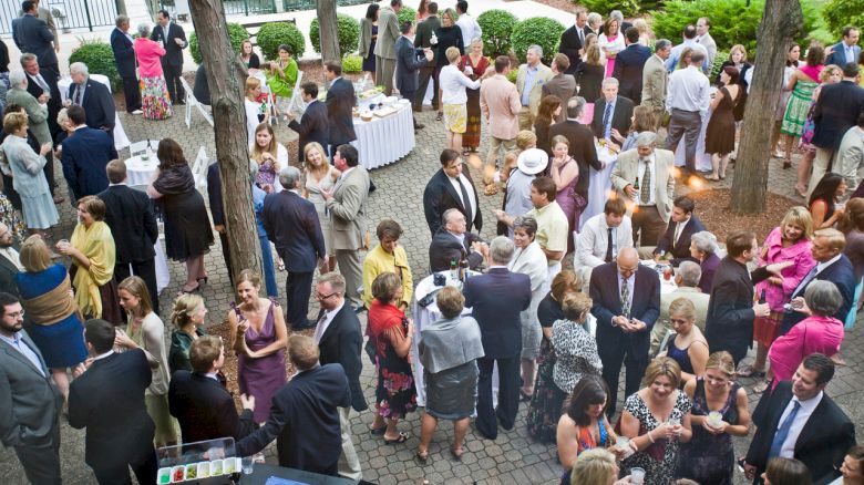 A large group of people are gathered at an outdoor event, mingling and socializing around tables and a drink station under trees.