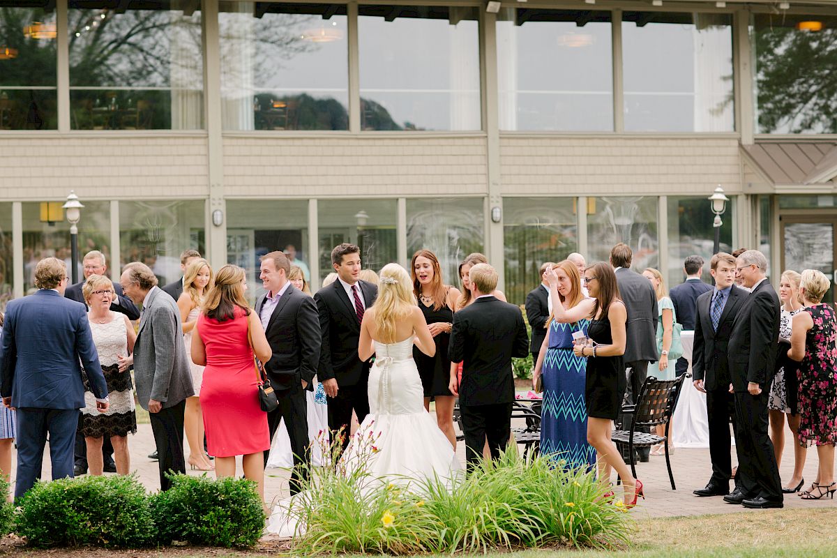 A group of people is gathered outdoors, many dressed formally, with a focus on a bride in a white dress, at what appears to be a wedding reception.