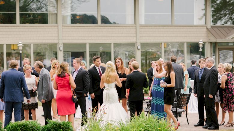 A group of people is gathered outdoors, many dressed formally, with a focus on a bride in a white dress, at what appears to be a wedding reception.