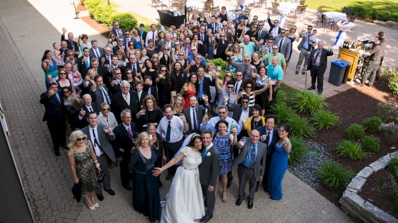 A large group of people, including a couple in wedding attire, are gathered outside for a group photo at what appears to be a wedding.