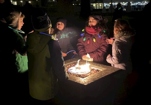 A group of children wearing winter clothing gather around a lit fire pit outdoors at night, illuminated by the fire’s light.
