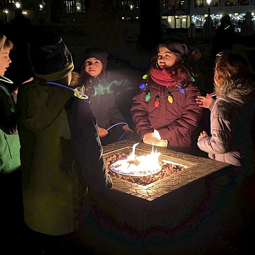 A group of children wearing winter clothing gather around a lit fire pit outdoors at night, illuminated by the fire’s light.