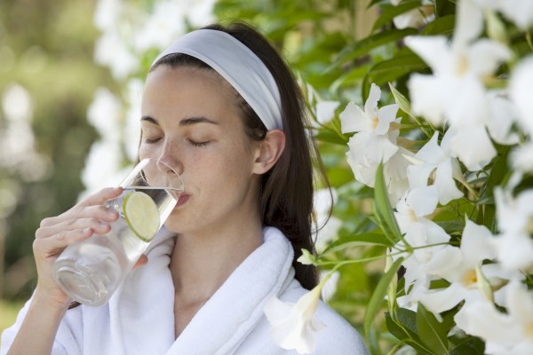 A woman with a headband and white towel drinks water with lemon slices outdoors among white flowers and greenery.