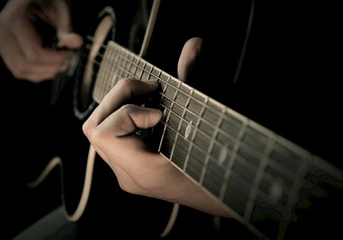 A close-up shot shows someone playing acoustic guitar with fingers on the fretboard and strumming with the other hand, captured in dim lighting.