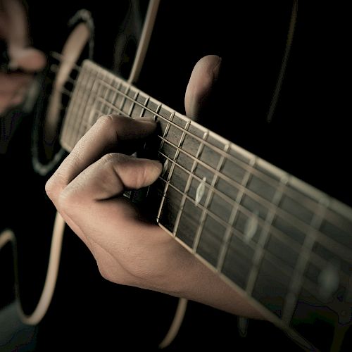 A close-up shot shows someone playing acoustic guitar with fingers on the fretboard and strumming with the other hand, captured in dim lighting.