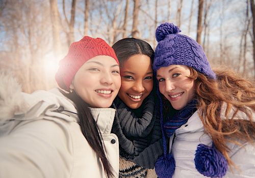 Three people wearing winter clothing are taking a selfie in a snowy forest, smiling and looking happy.