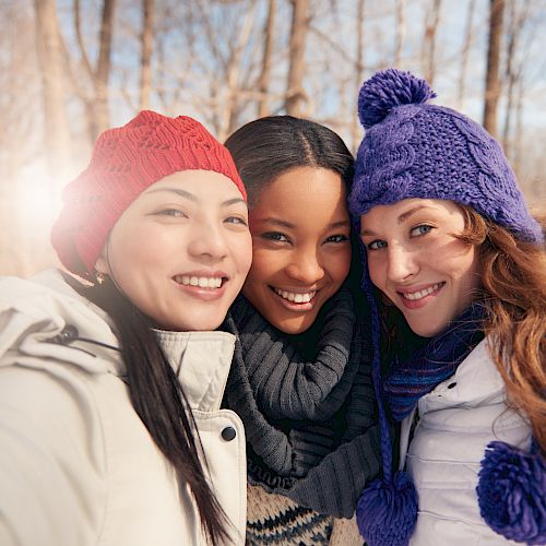 Three people wearing winter clothing are taking a selfie in a snowy forest, smiling and looking happy.